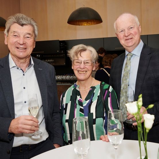 Drei Personen stehen zusammen an einem Tisch und halten Weingläser in der Hand. Der Mann auf der linken Seite lächelt. Die Frau in der Mitte trägt eine Brille und eine Halskette. Der Mann auf der rechten Seite trägt eine Krawatte. Hinter ihnen befindet sich ein Küchenbereich mit einigen Personen.