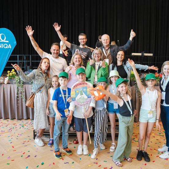 Eine Gruppe von Erwachsenen und Kindern posiert für ein Foto auf einer Bühne mit einem mit Konfetti bestreuten Boden, lächelt und hebt die Hände. Dahinter sind ein Banner mit dem Text 'QIQA' und ein schwarzer Hintergrund zu sehen.