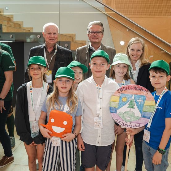 Eine Gruppe lächelnder Kinder und Erwachsener steht zusammen, trägt grüne Mützen und Abzeichen. Hinter ihnen stehen zwei Männer und eine Frau, wobei ein Kind ein Schild hält.