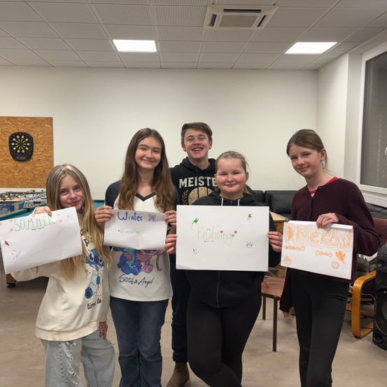 Fünf Mädchen halten Schilder mit den Jahreszeiten hoch und stehen in einem Raum mit weißen Wänden und Decken. Dahinter befindet sich ein Dartboard an der Wand und eine Couch.