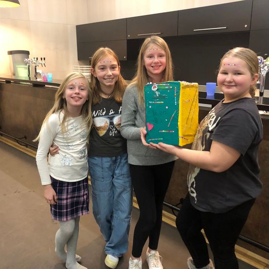 Vier junge Mädchen lächeln für ein Foto und halten eine dekorierte Box in der Hand, stehen in einer Küchenecke mit einer Arbeitsplatte, Schränken und einem Wasserhahn.