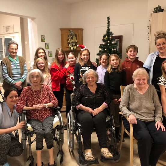 Eine Gruppe älterer Frauen in Rollstühlen und jüngerer Menschen versammelt sich zu einem Foto in einem festlichen Raum mit einem Weihnachtsbaum.