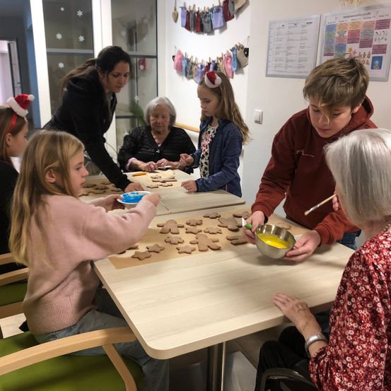 Eine Gruppe von Kindern und Erwachsenen sitzt um einen Tisch herum und macht Lebkuchen. Die Kinder tragen Weihnachtsmützen und haben Schüsseln mit Plätzchenteig. Eine erwachsene Frau hilft einem Jungen mit einem Pinsel.