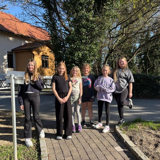 Eine Gruppe junger Mädchen steht auf einem gepflasterten Weg vor einem Haus mit rotem Dach und Zaun. Sie alle lächeln und posieren für ein Foto.