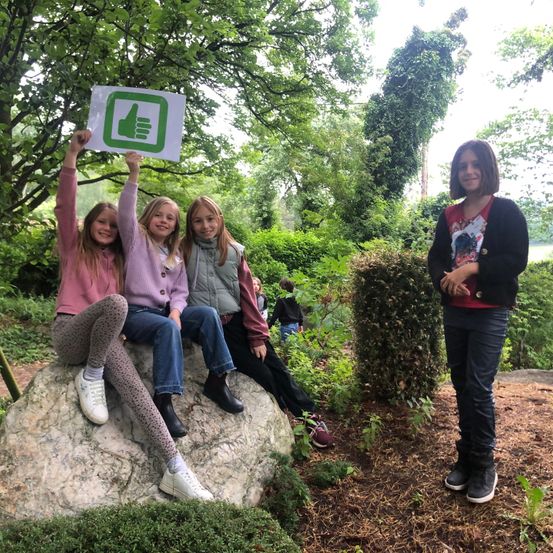 Vier Mädchen sind in einem Park. Drei sitzen auf einem Felsen und halten ein grünes Schild mit einem Daumen nach oben. Das vierte steht in der Nähe.