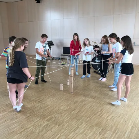 Eine Gruppe von Kindern versammelt sich in einem Raum und hält an Schnüren befestigte Holzblöcke, wahrscheinlich in einer Teamaktivität engagiert.