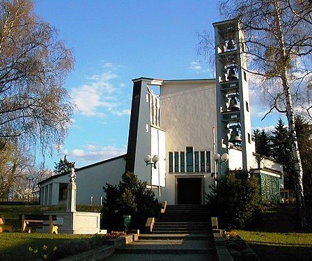 Eine moderne weiße Kirche mit Turm und Glocken. Sie ist von Bäumen umgeben und hat eine Bank und eine Statue davor.