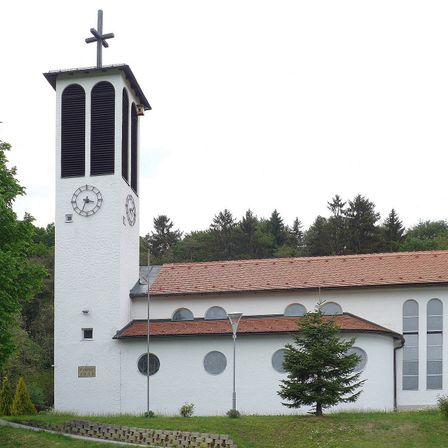 Eine weiße Kirche mit einem roten Dach und einem hohen Turm mit einem Kreuz oben. Es hat bogenförmige Fenster und eine Uhr auf dem Turm.