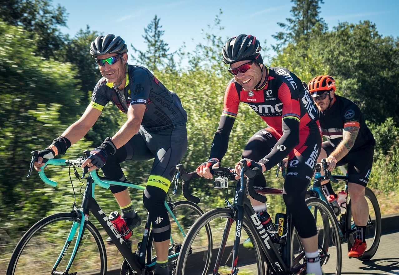 Drei Radfahrer in Radsportbekleidung fahren auf einer von Bäumen gesäumten Straße. Die Radfahrer tragen Helme und Sonnenbrillen. Der Mann vorne hat eine Wasserflasche an seinem Fahrrad befestigt.