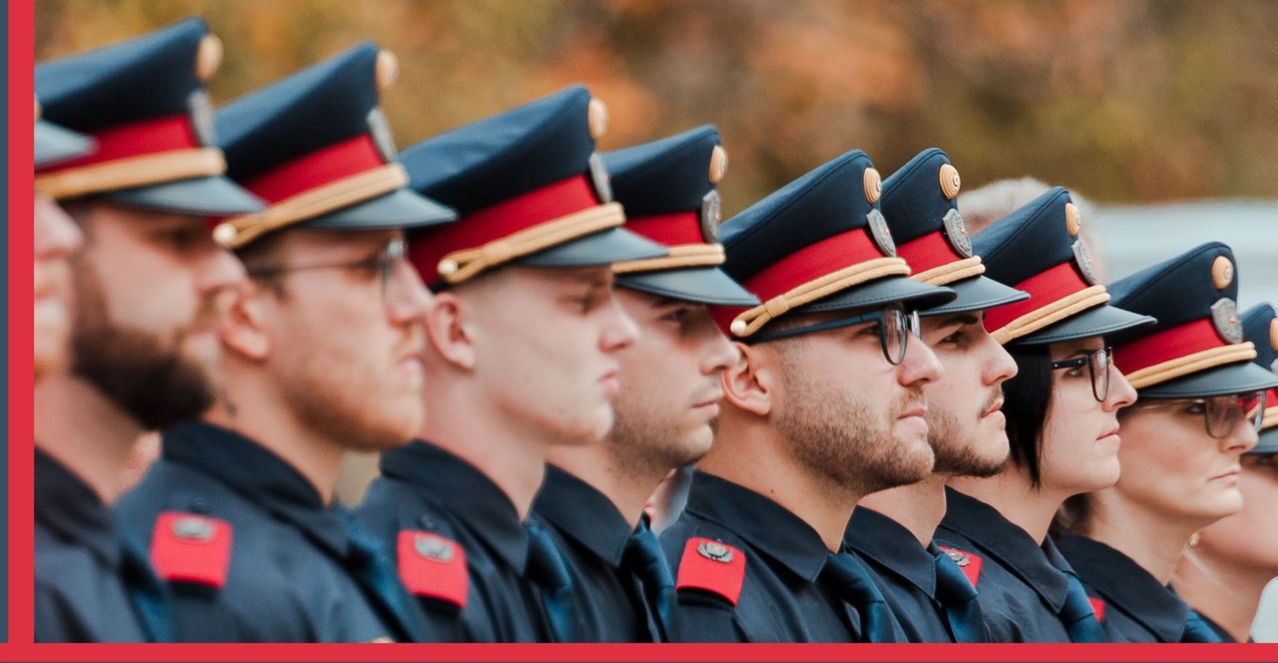 Fünf uniformierte Offiziere stehen in einer Reihe und tragen Mützen mit roten und blauen Akzenten. Sie schauen geradeaus, möglicherweise bei einer Parade.