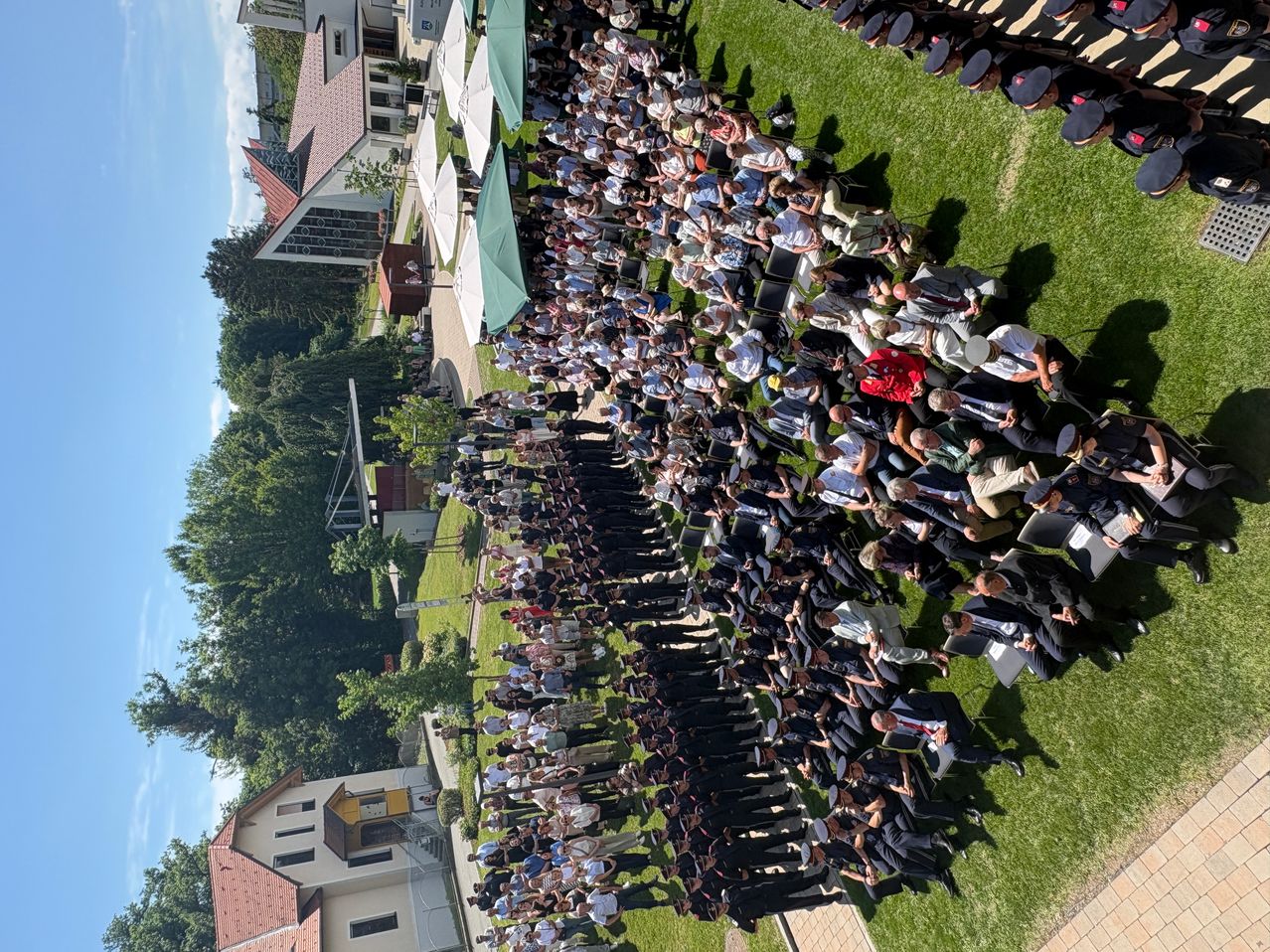 Eine große Menschenmenge, einige in Uniformen, versammelt sich auf einem Rasenfeld für eine Veranstaltung. Gebäude und Bäume im Hintergrund.