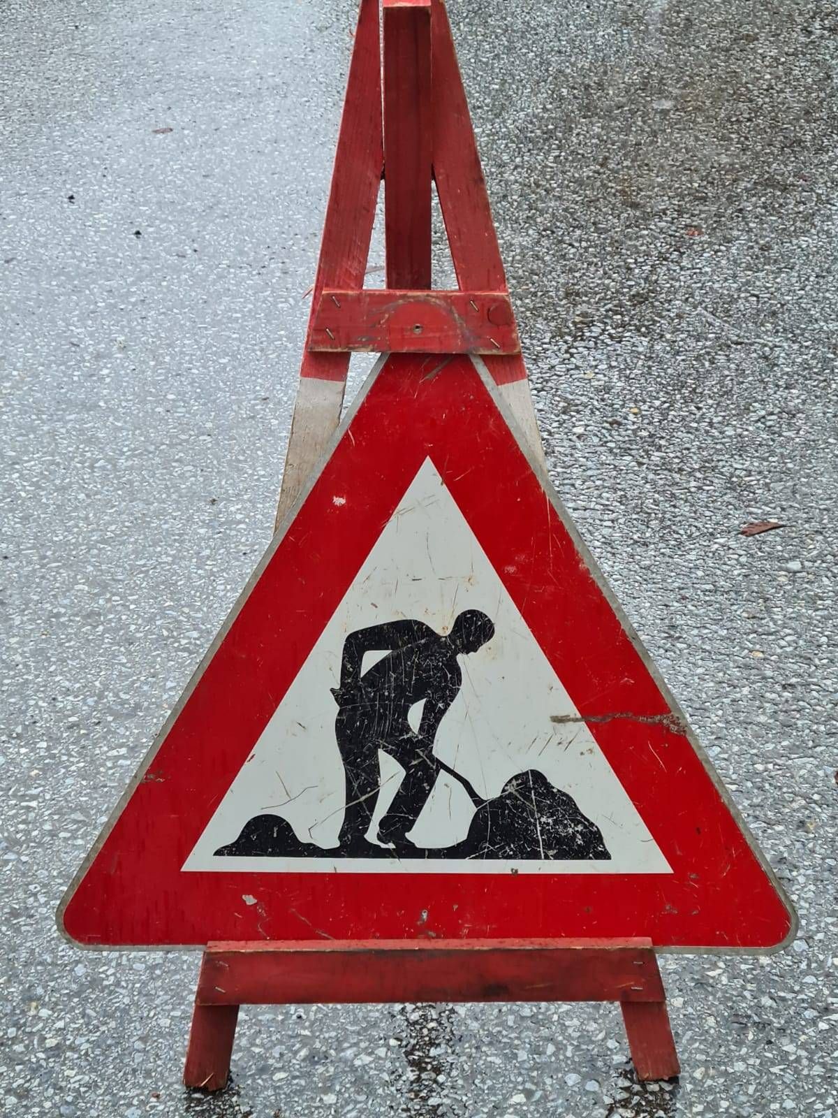 Ein dreieckiges Verkehrsschild mit rotem Rand, das eine Silhouette einer Person beim Graben zeigt, auf einer Kiesoberfläche platziert.