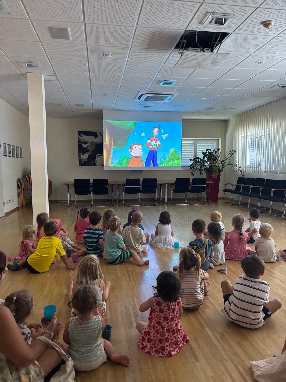 Eine Gruppe von Kindern sitzt auf dem Boden und schaut einen Cartoon auf einer Projektionsleinwand in einem Raum mit Holzböden und weißen Wänden.
