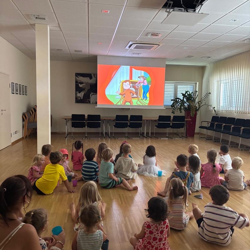 Eine Gruppe von Kindern sitzt in einem Raum und schaut sich einen Cartoon auf einer Projektionsleinwand an. Der Raum ist gut beleuchtet mit Holzböden und Stühlen, die in einem Kreis angeordnet sind.