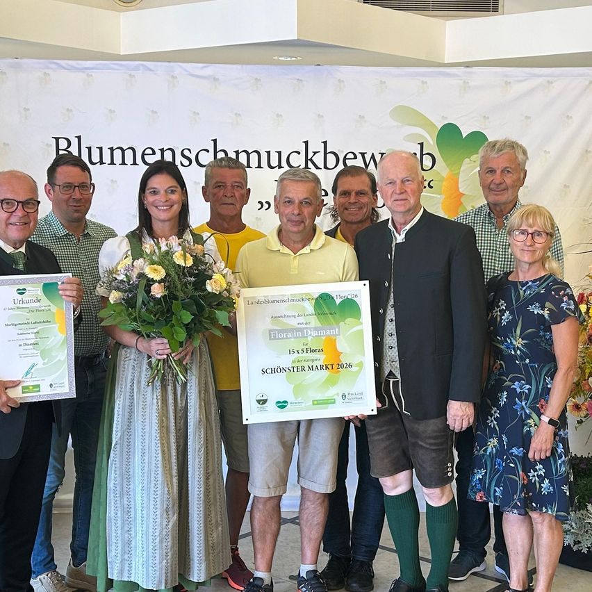 A group of people are posing for a photo with certificates and flowers. A woman in the middle holds a bouquet of flowers. Behind them, a banner reads 'Blumenschmuckbewerb'.