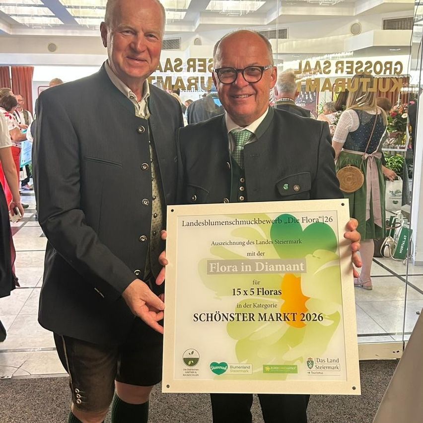 Two men in traditional attire hold a plaque with a floral design, smiling for the camera. The plaque reads 'Flora in Diamant' and 'Schonster Markt 2026'. Behind them, a glass wall reflects a bustling indoor market.
