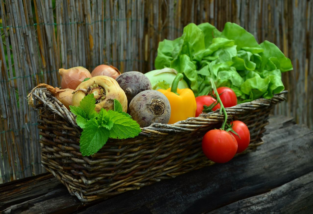 A wicker basket filled with fresh vegetables including onions, peppers, and tomatoes, along with green leaves and herbs.