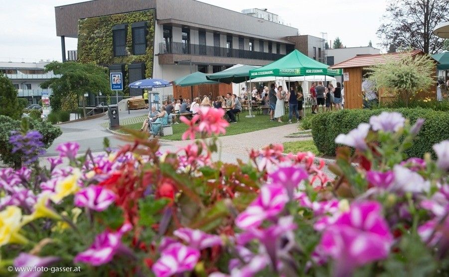 A vibrant outdoor gathering with people seated under green tents, surrounded by colorful flowers and a modern building with a green wall.