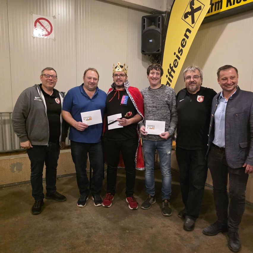 Fünf Männer posieren für ein Foto in einem Raum und halten Urkunden in der Hand. Der Mann in der Mitte trägt eine Krone und einen Umhang. Hinter ihnen befindet sich ein gelber Banner.