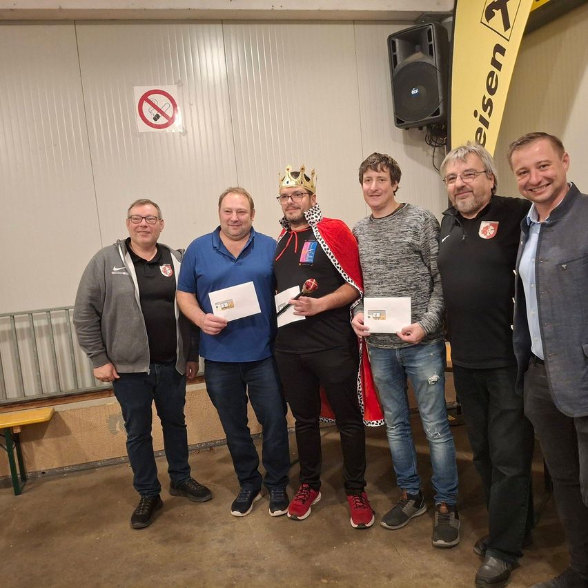 Fünf Männer stehen drinnen zusammen. Der Mann in der Mitte trägt eine Krone und einen roten Umhang. Sie alle halten Urkunden und lächeln. Im Hintergrund befinden sich ein Rauchverbotsschild und ein gelber Banner.