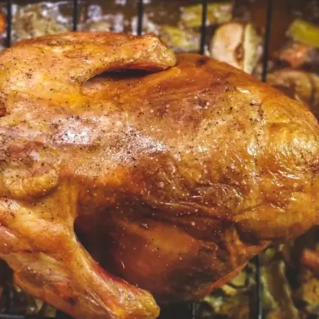 Ein gebratenes Huhn befindet sich auf einer Metallschale mit Gemüse im Hintergrund. Das Huhn hat eine knusprige Haut und eine goldbraune Farbe. Es liegt auf einem Grillrost.