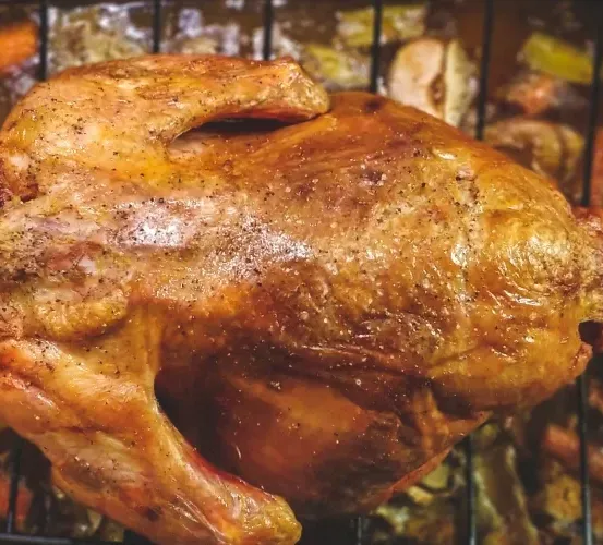 Ein gebratenes Huhn befindet sich auf einer Metallschale mit Gemüse im Hintergrund. Das Huhn hat eine knusprige Haut und eine goldbraune Farbe. Es liegt auf einem Grillrost.
