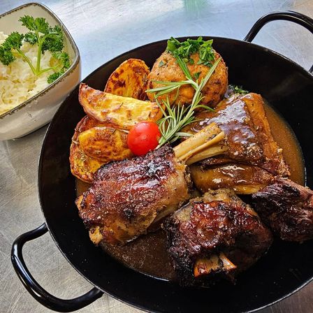 Eine schwarze Pfanne enthält gebratenes Lamm mit Sauce, garniert mit Rosmarin, serviert mit gebratenen Kartoffeln und einer Schüssel Reis.