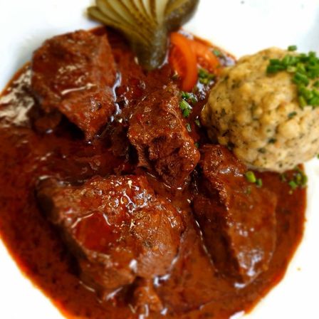 Ein Gericht aus geschmortem Rindfleisch in reicher Sauce, begleitet von Kartoffelpüree und Essiggurken, wird auf einem weißen Teller serviert.