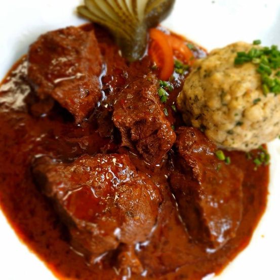 Ein Gericht aus geschmortem Rindfleisch in reicher Sauce, begleitet von Kartoffelpüree und Essiggurken, wird auf einem weißen Teller serviert.