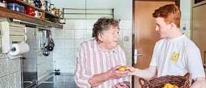 Eine ältere Frau, die eine Zitrone hält, steht in einer Küche und lächelt einen Mann an, der einen Korb mit Zitronen hält. Die Küche hat geflieste Wände, eine Spüle und Utensilien auf einem Regal.