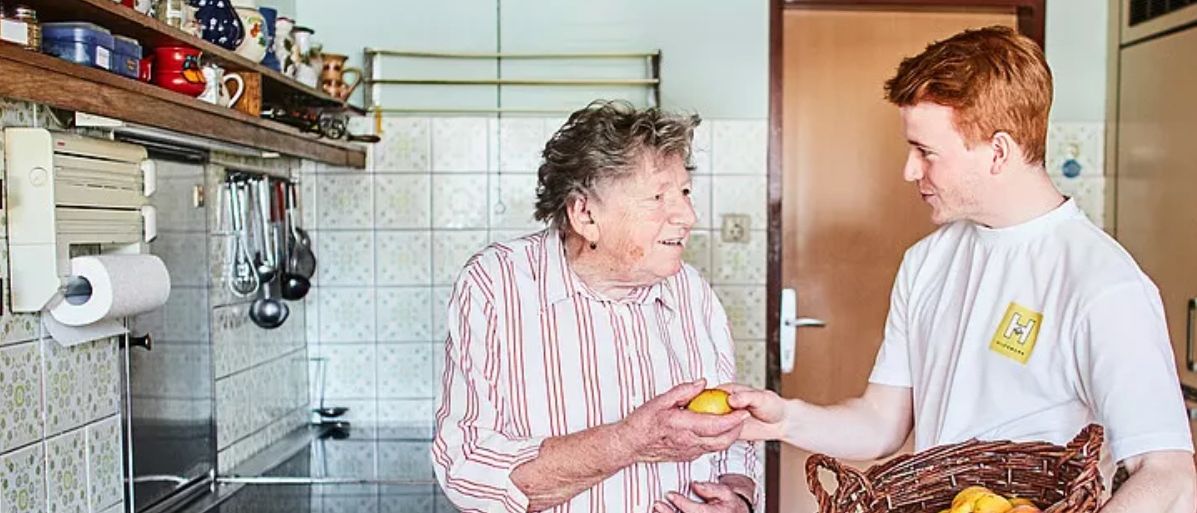 Eine ältere Frau, die eine Zitrone hält, steht in einer Küche und lächelt einen Mann an, der einen Korb mit Zitronen hält. Die Küche hat geflieste Wände, eine Spüle und Utensilien auf einem Regal.