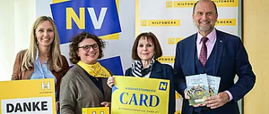 Fünf Personen posieren für ein Foto vor einem Banner. Das Banner trägt in großen Buchstaben 'NV'. Die Frau auf der linken Seite hält ein Schild mit der Aufschrift 'DANKE'. Der Mann auf der rechten Seite hält ein Buch und ein Schild mit der Aufschrift 'CARD'.