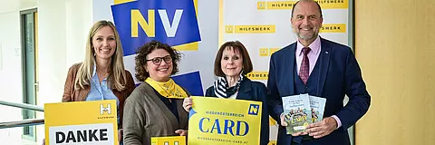 Fünf Personen posieren für ein Foto vor einem Banner. Das Banner trägt in großen Buchstaben 'NV'. Die Frau auf der linken Seite hält ein Schild mit der Aufschrift 'DANKE'. Der Mann auf der rechten Seite hält ein Buch und ein Schild mit der Aufschrift 'CARD'.