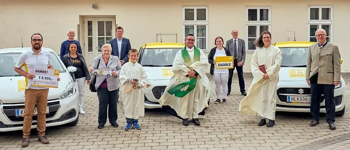 Eine Gruppe von Menschen steht vor zwei Autos, einem gelben und einem weißen, vor einem Gebäude. Sie sind in Roben gekleidet und halten Gegenstände. Ein Priester steht zwischen den Autos.