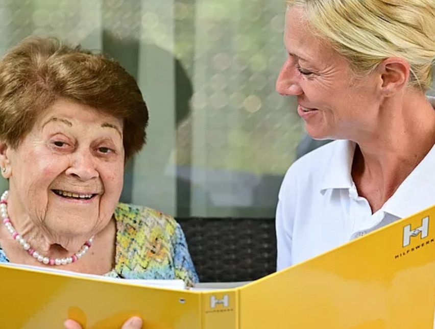 Eine ältere Frau lächelt, während sie ein gelbes Buch liest, neben ihr steht eine junge Frau. Die junge Frau trägt ein weißes Poloshirt und beide schauen auf den Inhalt des Buches.