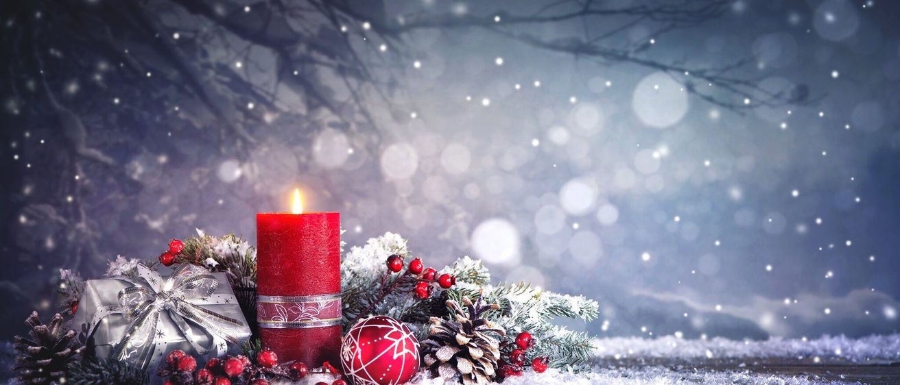 Eine rote Kerze, umgeben von Weihnachtsdekorationen wie Tannenzapfen, Stechbeeren und einer roten Kugel, mit einem verschwommenen verschneiten Hintergrund.