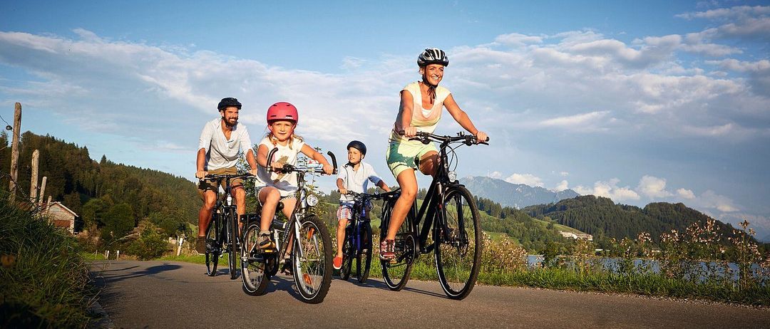 Eine Familie fährt auf einer Straße mit Bergen im Hintergrund mit Fahrrädern. Die Mutter und die Kinder tragen Helme. Der Vater trägt einen Helm und hält das Lenkrad.