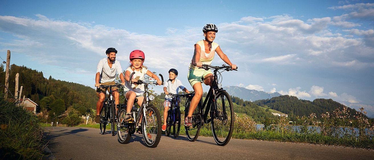 Eine Familie fährt auf einer Straße mit Bergen im Hintergrund mit Fahrrädern. Die Mutter und die Kinder tragen Helme. Der Vater trägt einen Helm und hält das Lenkrad.