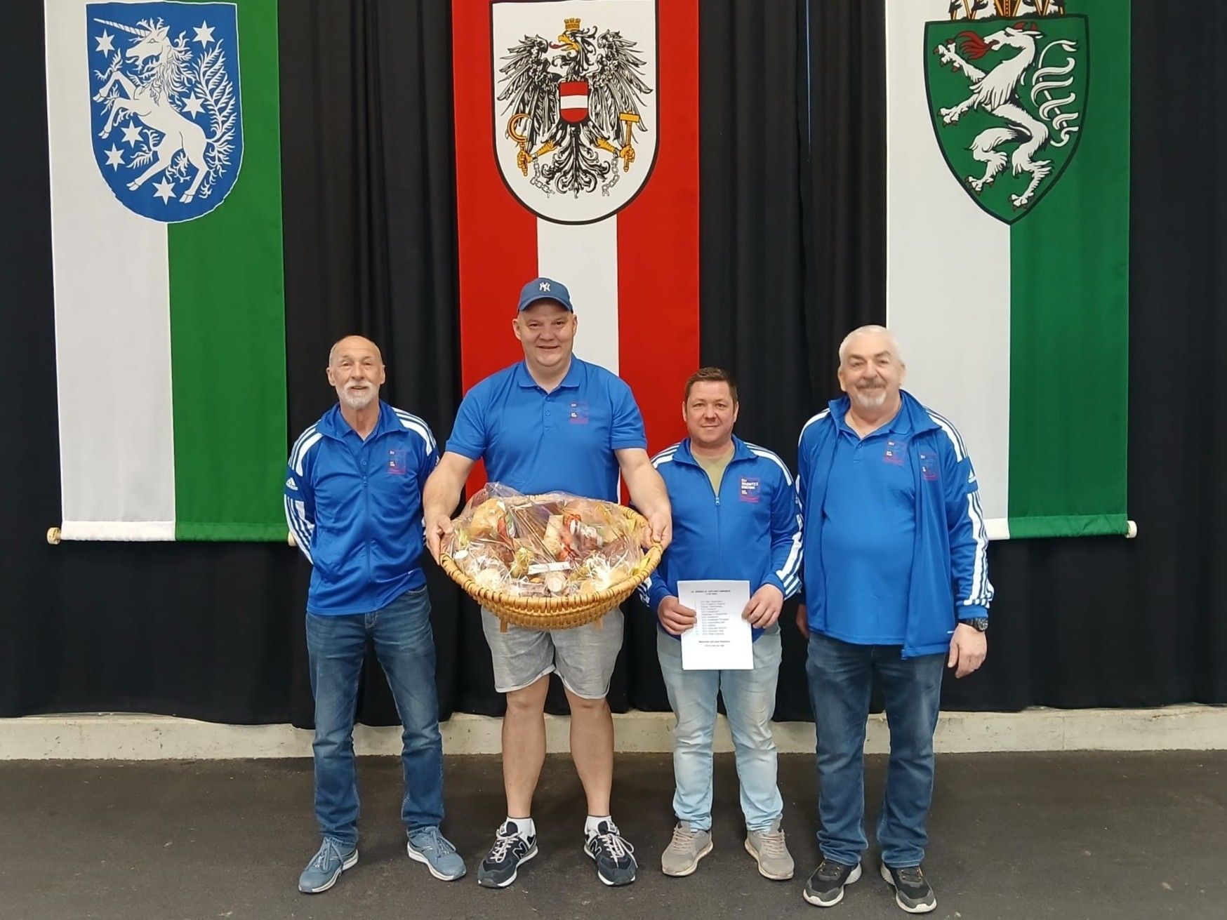 Vier Männer stehen vor einer Wand mit drei verschiedenen Flaggen. Ein Mann hält einen Korb mit Essen.