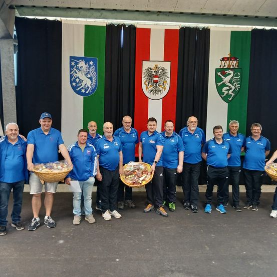 Eine Gruppe von Männern in blauen Hemden posiert für ein Foto mit Körben voller Essen. Dahinter hängen Flaggen und Wappen.