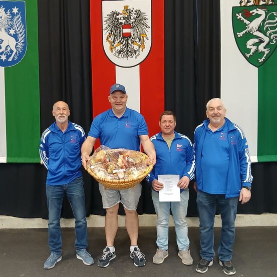 Vier Männer in blauen Jacken und Mützen posieren für ein Foto vor einem Hintergrund mit österreichischen Flaggen und Wappen. Der Mann in der Mitte hält einen Korb voller Essen.