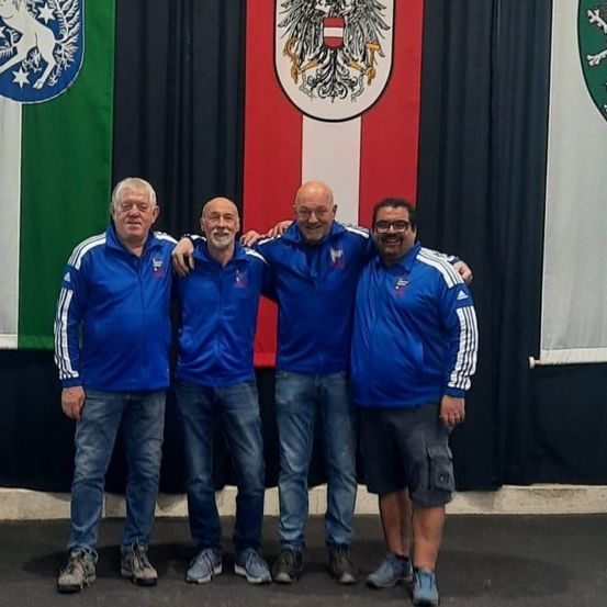 Vier Männer posieren für ein Foto vor drei Flaggen. Die Flaggen sind grün, rot und weiß und haben verschiedene Symbole darauf. Die Männer tragen blaue Jacken und Jeans.