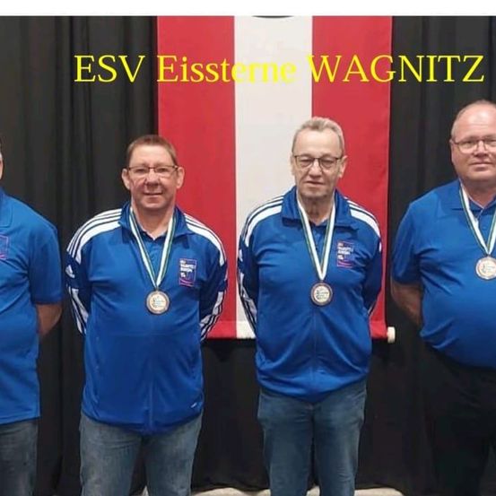 Vier Männer in blauen Hemden und mit Brillen stehen zusammen, möglicherweise bei einer Preisverleihung. Sie tragen Medaillen um den Hals. Hinter ihnen ist ein Hintergrund mit einem rot-weiß gestreiften Muster.