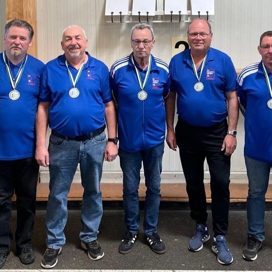 Fünf Männer stehen nebeneinander und tragen blaue Hemden und Jeans. Sie haben Medaillen um den Hals. Sie tragen alle Turnschuhe und lächeln.