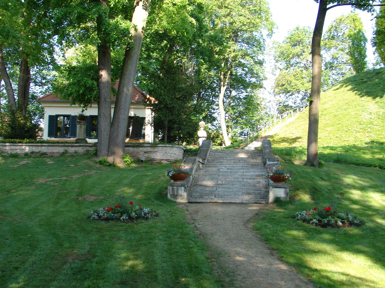Ein kleines Haus mit einem Steinweg, der zu einer Steintreppe führt. Die Treppe hat eine Statue oben. Auf beiden Seiten des Weges befinden sich Pflanzen und Blumen.