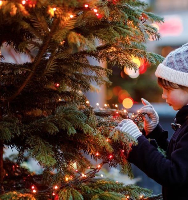 Ein junges Kind schmückt einen Weihnachtsbaum mit roten Lichtern, trägt einen Wintermütze und Handschuhe.