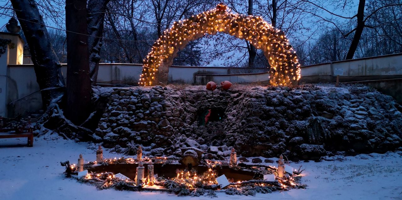 A snow-covered area features a lit archway with hanging lights and two pumpkins. Additional lights and decorative items are scattered around. Trees and a wall are visible in the background.