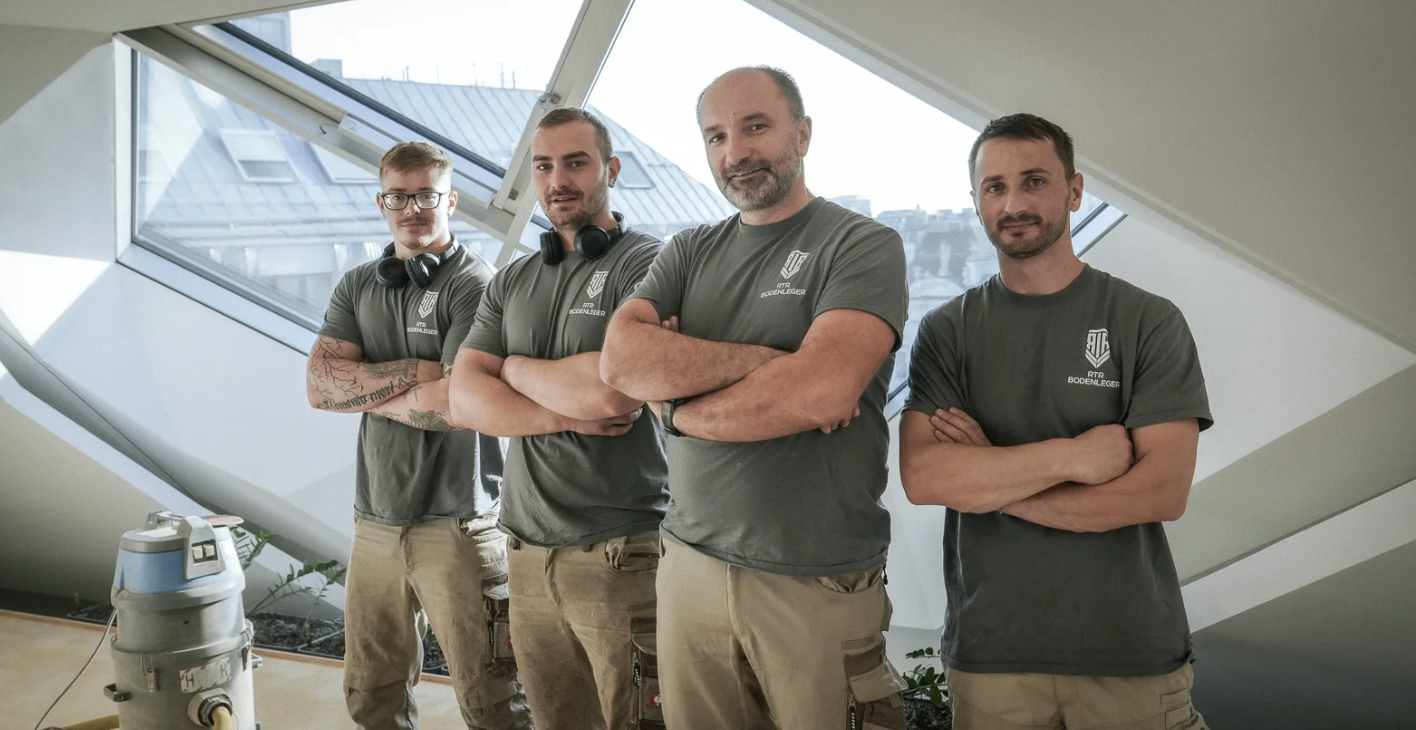 Vier Männer in grauen Arbeitshemden mit Logos stehen nebeneinander, lächeln für die Kamera mit verschränkten Armen. Dahinter befindet sich ein großes Fenster mit Blick auf eine Stadtlandschaft.
