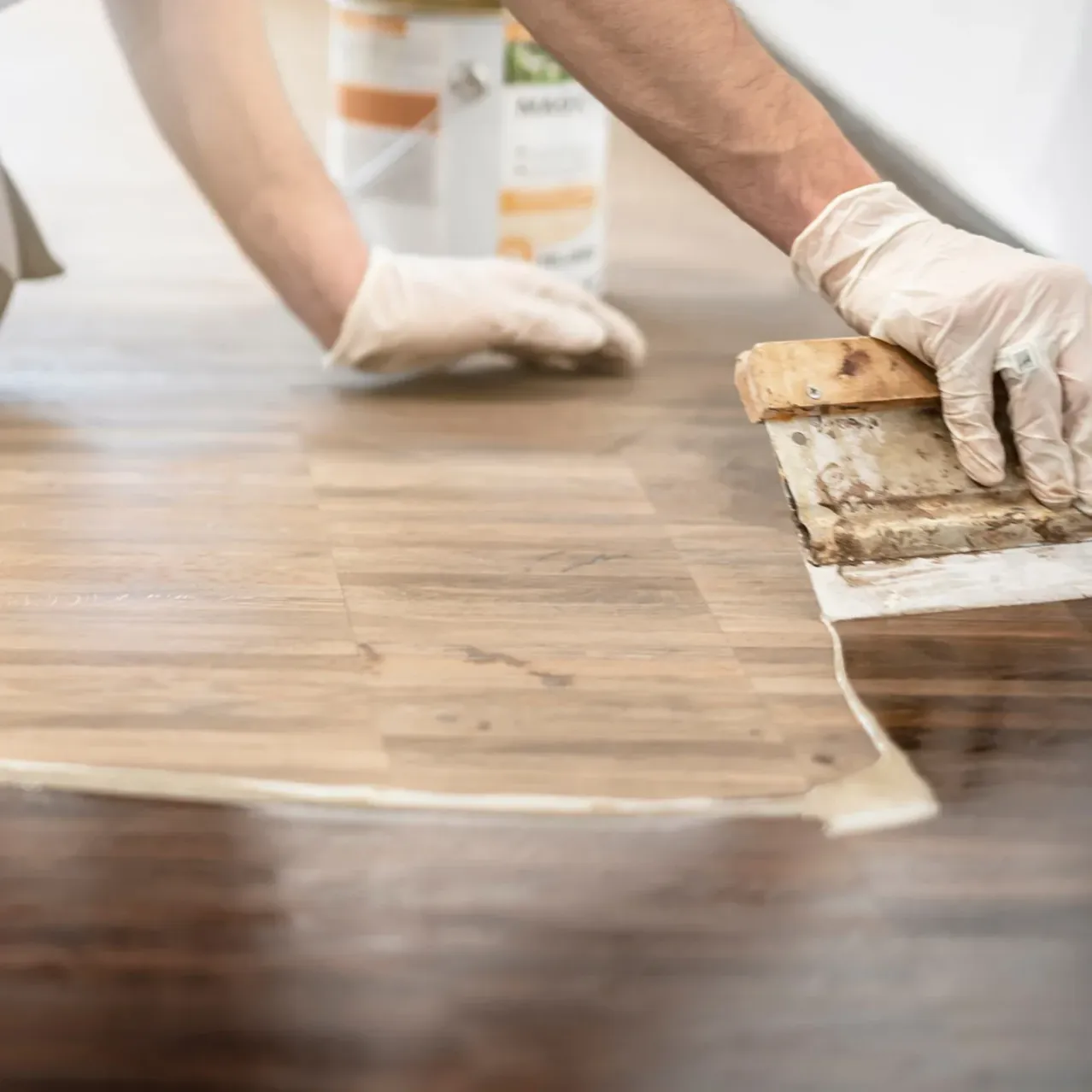 Eine Person mit Handschuhen trägt eine Substanz auf einen Holzboden auf, wahrscheinlich ein Versiegelungs- oder Finishmittel. Der Boden ist sauber und scheint gut gepflegt zu sein.