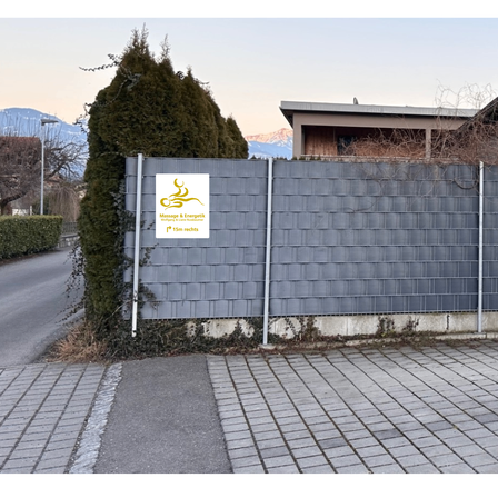 Ein grauer Holzzaun mit Logo und dem Text Massage & Energietechnik steht vor einem Gebäude mit Bergen im Hintergrund.